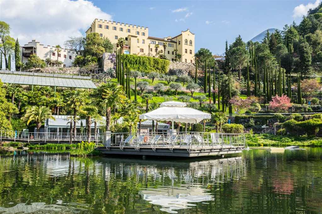 Scenic view of the Gardens Trauttmannsdorf overlooking a reflective lake, surrounded by lush gardens and palm trees.