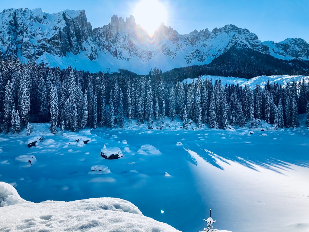 A snowy landscape featuring tall evergreen trees, a frozen lake, and majestic Dolomite mountains in the background under a clear blue sky with a bright sun.