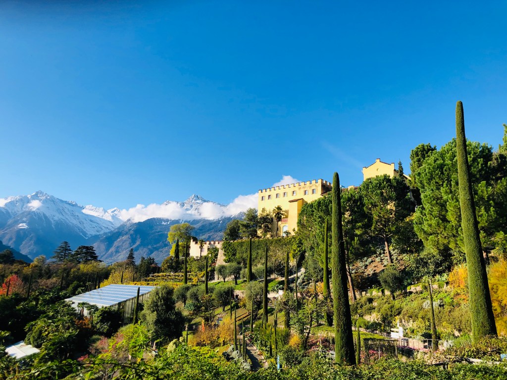 A scenic view of a castle surrounded by tall cypress trees and lush gardens, with snow-capped mountains in the background under a clear blue sky.